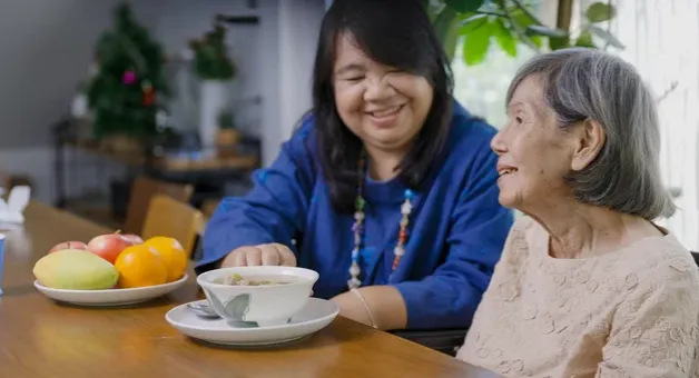A caregiver and an older woman sharing tea and fruit at a table