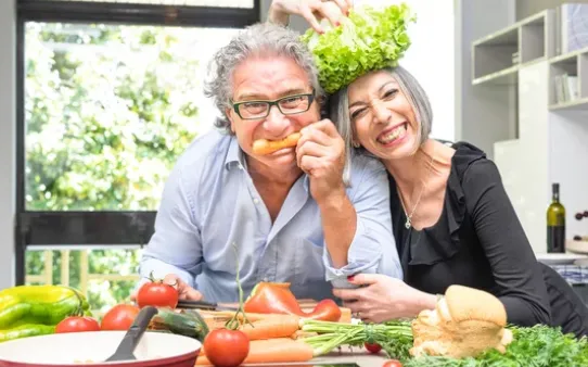 An older couple laughing while cooking with fresh vegetables