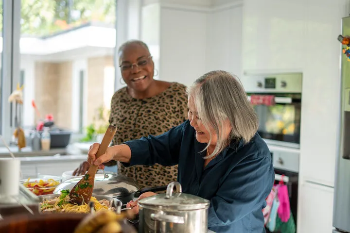 Two older women laughing while cooking pasta together in a home kitchen
